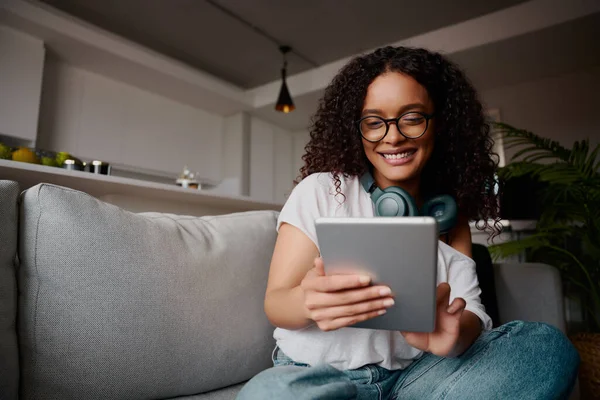 Multi-culturele jonge volwassen vrouw met tablet op de bank. Glimlachend met bril op — Stockfoto