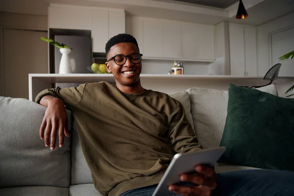 Jovem adulto preto macho sorrindo para tablet enquanto sentado no salão no sofá — Fotografia de Stock