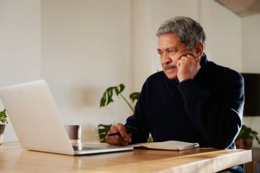 Dizüstü bilgisayardan haber okuyan çok kültürlü yaşlı bir erkek. Modern evde izole edilmiş, mutfak tezgahında oturuyor..