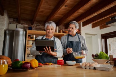 Modern mutfakta yemek tarifi için tablet kullanan çok kültürlü yaşlı bir çift. Mutlu çift sağlıklı yemek pişiriyor..