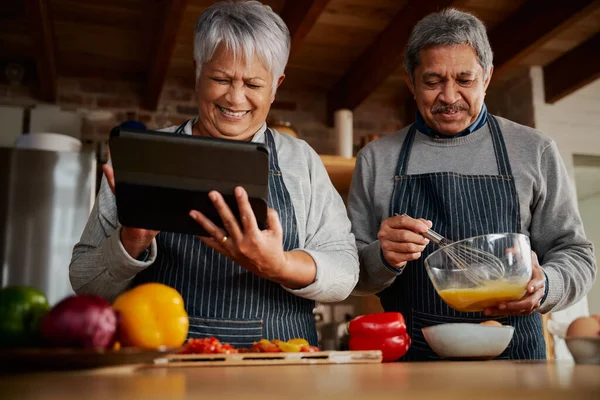 Modern mutfakta kocasıyla yemek tarifi araştırmak için tablet kullanan çok kültürlü yaşlı bir kadın. Mutlu çift sağlıklı yemek pişiriyor..