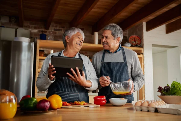 Modern mutfakta kahvaltı hazırlarken birlikte gülen çok kültürlü yaşlı bir çift sağlıklı yemek tarifi için tablet kullanıyor..
