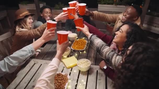 Diverso grupo de amigos brindando entre sí mientras se sientan alrededor de una mesa en una fiesta en la azotea celebrando — Vídeo de stock