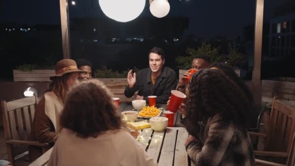 Grupo multiétnico de amigos adultos que socializan alrededor de una mesa en una fiesta en la azotea de la ciudad — Vídeos de Stock