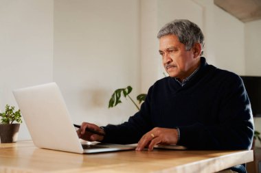 Dizüstü bilgisayarda yazan çok kültürlü yaşlı bir erkek. Modern evde izole edilmiş, mutfak tezgahında oturuyor..