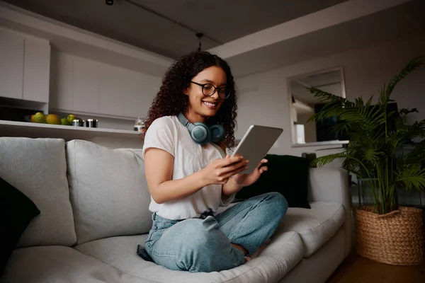 Modern apartmandaki kanepede tablet kullanırken çok kültürlü bir kadın gülümsüyor. — Stok fotoğraf