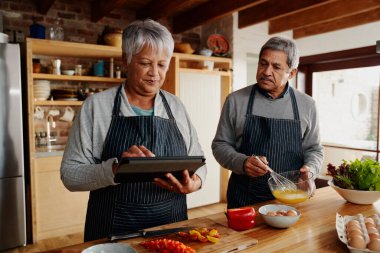 Kocasıyla birlikte modern mutfaklarında kahvaltı tarifini uygulamak için elektronik tablet kullanan çok kültürlü yaşlı bir kadın..