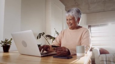 Çok kültürlü yaşlı bir kadın dizüstü bilgisayardan online alışveriş yapıyor. Mutfak tezgahında kredi kartıyla oturmak.