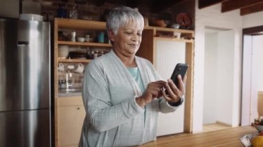 Çok kültürlü, yaşlı ve akıllı telefonuyla yazan emekli bir kadın. Mutlulukla modern mutfakta duruyorum.