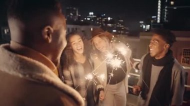 Gece vakti çatıdaki bir partide havai fişeklerini yakarken gülen bir grup arkadaş..
