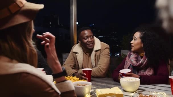 Hombre negro charlando con sus amigos multiétnicos y sentado en una mesa en una fiesta en la azotea de la ciudad por la noche — Vídeo de stock