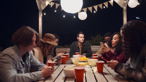 Одинокий белый мужчина, сидящий за столом, в то время как его разнообразная группа друзей разговаривает по телефону на вечеринке. — стоковое видео