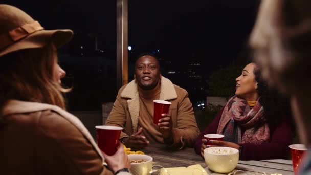 Черный мужчина произносит тост со своими многонациональными друзьями и сидит за столом на вечеринке на крыше города в ночное время — стоковое видео