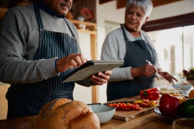 Çok kültürlü yaşlı bir çift mutfakta yemek tarifi için tablet kullanıyor. Mutlu bir emeklilik sağlıklı yemek pişirmek..
