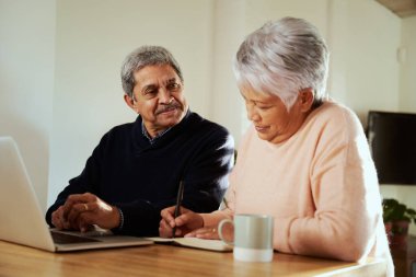 Modern mutfak tezgahında dizüstü bilgisayarlarıyla oturan çok kültürlü yaşlı bir çift. Karısı not defterine not tutuyor. Emeklilik bütçesi.