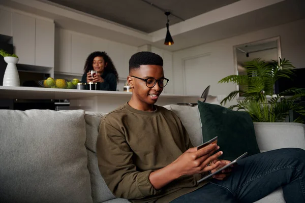 Noir mâle faire des achats en ligne sur canapé tandis que la petite amie est dans la cuisine, détendu et heureux — Photo