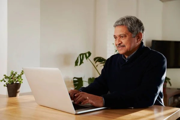 Мультикультурный пожилой мужчина переписывается по электронной почте при помощи ноутбука. Изоляция в современном доме, сидя за кухонным столиком. — стоковое фото