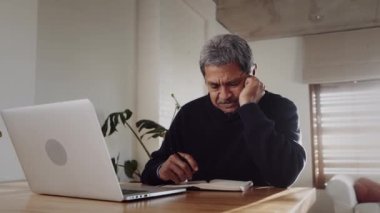 Çok kültürlü yaşlı erkek, dizüstü bilgisayar kullanıyor ve kalem ve kağıtla notlar alıyor. Mutfak tezgahında oturuyor.