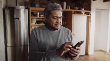 Çok kültürlü, yaşlı bir erkek akıllı telefonda yazı yazıyor. Modern mutfakta duruyor.