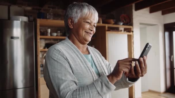 Akıllı telefonda yazarken gülümseyen çok kültürlü yaşlı bir kadın. Modern mutfakta duruyor. — Stok video