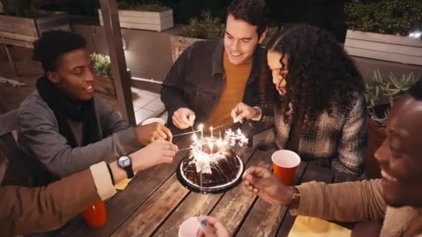Różnorodna grupa przyjaciół zapala ognie wokół tortu urodzinowego. Siedząc wokół stołu na dachu w nocy. — Wideo stockowe