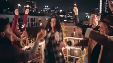 Çok kültürlü bir grup yetişkin arkadaş çatıda kıvılcımlarla dans ediyor ve arkalarında şehir var..