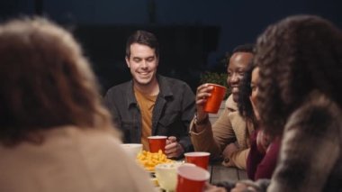 Beyaz erkek, şehirdeki bir çatı partisinde çok ırklı bir grup arkadaşıyla masanın etrafında sosyalleşiyor.