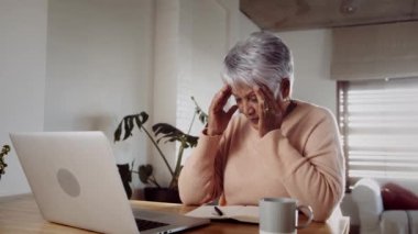 Çok kültürlü yaşlı bir kadın üzgün, dizüstü bilgisayarında kötü haberler alıyor, mutfak tezgahında oturuyor.
