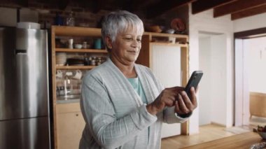 Akıllı telefon ekranında yazan mutlu çok kültürlü yaşlı kadın. Modern mutfakta duruyor.