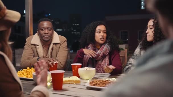 Multikulturní žena vypráví příběh své rozmanité skupině přátel, kteří sedí u stolu na střešní párty. — Stock video
