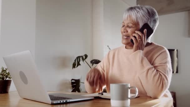 Multikulturelle Seniorin glücklich am Telefon, wenn sie mit Laptop und Notizbuch am Küchentisch sitzt — Stockvideo
