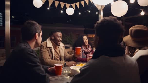 Siyah erkek, gecenin bir yarısı dışarıda bir masada, çok ırklı yetişkin arkadaş grubunun şerefine kadeh kaldırmak için ayakta duruyor. — Stok video