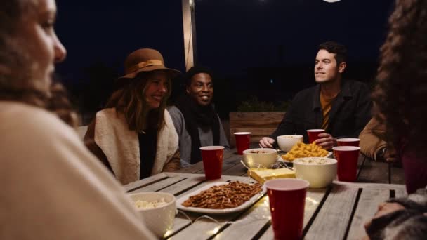 Різні дорослі групи друзів спілкуються і сміються на вечірці на відкритому повітрі за столом в нічний час — стокове відео
