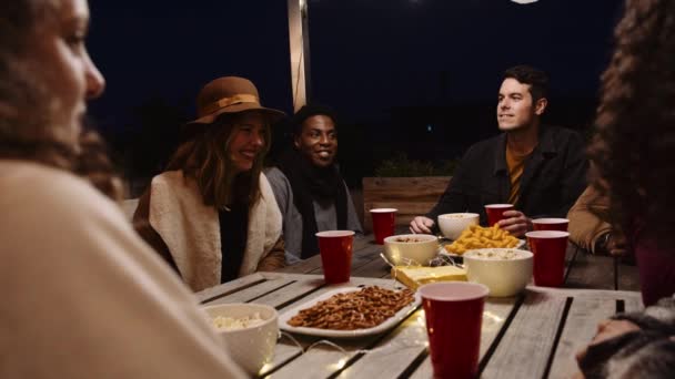 Diverse adult group of friends chatting and laughing at a party outdoors at a table in the night time — 图库视频影像