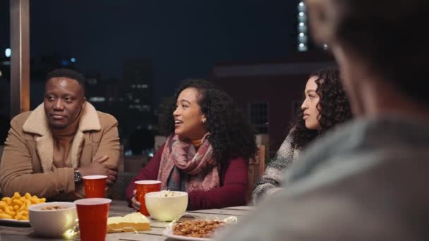 Multi-kulturel kvindelig fortæller en historie omkring et bord til hendes forskelligartede gruppe af venner på et tag fest. – Stock-video