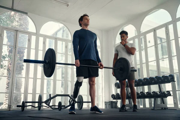 Паралимпийский спортсмен тяжелой атлетики со своим тренером в спортзале. Человек с протезной ногой тренируется у своего инструктора — стоковое фото
