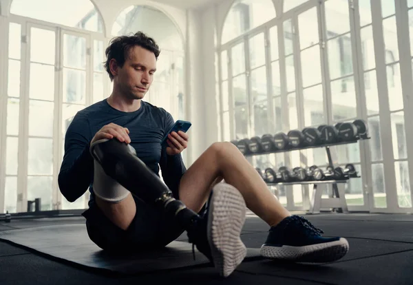 Paralympische atleet op zijn mobiel tijdens de gymnastiektraining. Man met een kunstbeen op zijn mobiele telefoon in een moderne fitnessruimte — Stockfoto