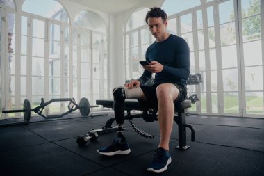 Jimnastik antrenmanında cep telefonuyla felçli bir sporcu. Protezli bir adam bankta oturmuş cep telefonuna bakıyor. .