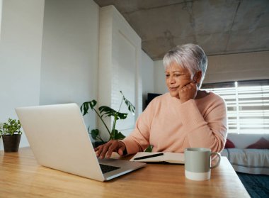 Dizüstü bilgisayarında interneti kaydırırken ilgili çok ırklı yaşlı bir kadın. Evdeki mutfak tezgahında oturmak..