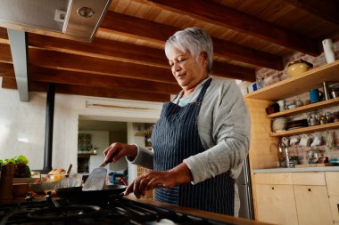 Modern mutfağındaki bir tavada kahvaltı hazırlayan çok kültürlü yaşlı kadın. Mutlu bir emeklilik, sağlıklı bir yaşam tarzı.