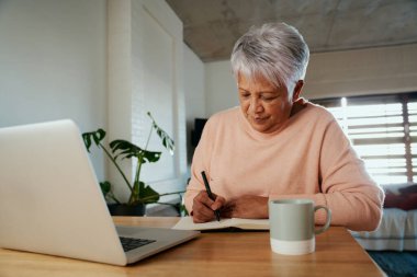Modern bankoda dizüstü bilgisayarın önünde otururken kalemle yazan çok ırklı yaşlı bir kadın..