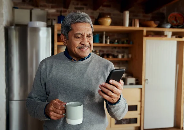 Счастливый многокультурный пожилой мужчина печатает сообщение на смартфоне, стоя на современной кухне, держа утренний кофе. — стоковое фото