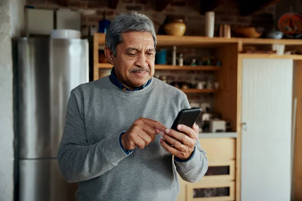 Содержание мультикультурного пожилого мужчины, печатающего сообщение на смартфоне, стоя на современной кухне. — стоковое фото