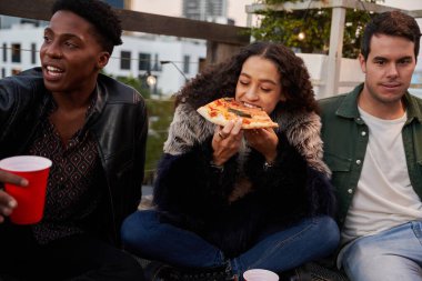 Bir grup çok kültürlü arkadaş terasta oturmuş lezzetli pizzalar yiyorlar. Çift ırklı dişiler yiyecekleri ısırıyor..