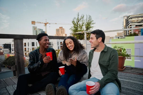 Grupo de jóvenes adultos multiculturales que se ríen de la broma juntos. Sentado en una fiesta en la azotea de la ciudad, buenos tiempos. — Foto de Stock