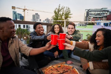 Bir grup farklı arkadaş şehirdeki terasta otururken kırmızı bardaklarla kadeh kaldırıyor.