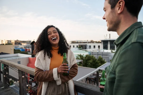 Biracial hembra adulta joven riendo con un amigo en el balcón de una terraza en la azotea de la ciudad. Bebida en la mano, vistas al paisaje urbano — Foto de Stock