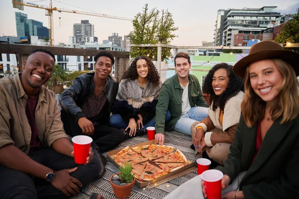 Şehirdeki bir terasta otururken kameraya kırmızı bardaklarla gülümseyen bir grup arkadaş.