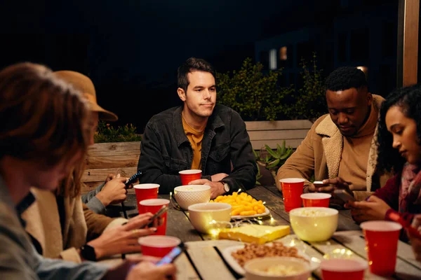 Biały mężczyzna znudzony, siedzący przy stole na imprezie, podczas gdy wszyscy jego przyjaciele smsują na telefonach. Technologia, media społecznościowe powodujące zachowania antyspołeczne — Zdjęcie stockowe