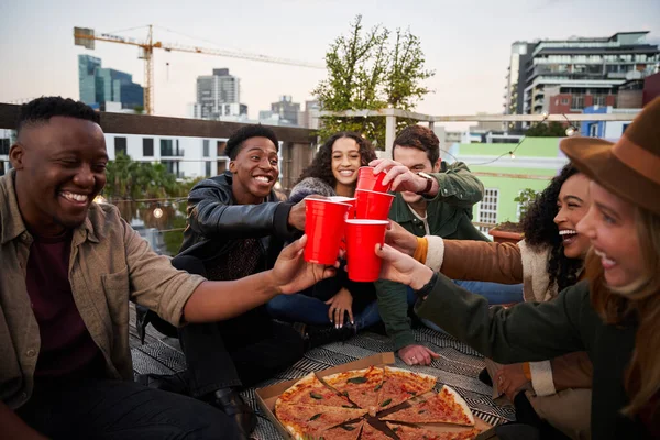 Группа разнообразных друзей произносит тост с красными чашками, сидя на террасе на крыше в городе. Хорошие времена и счастье — стоковое фото